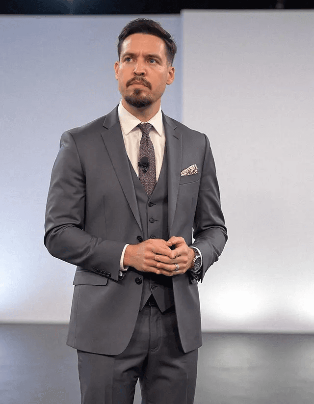 Adrian Arribas wearing a grey three-piece suit with a purple tie in a professional studio setting